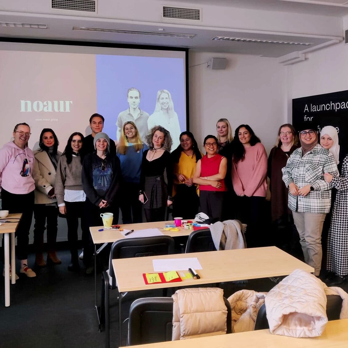 Group photo of a diverse team standing in a classroom setting, with a presentation slide in the background displaying the 'Noaur' logo and website. The slide features two individuals' portraits side by side. Tables in the foreground are scattered with papers, cups, and office supplies.
